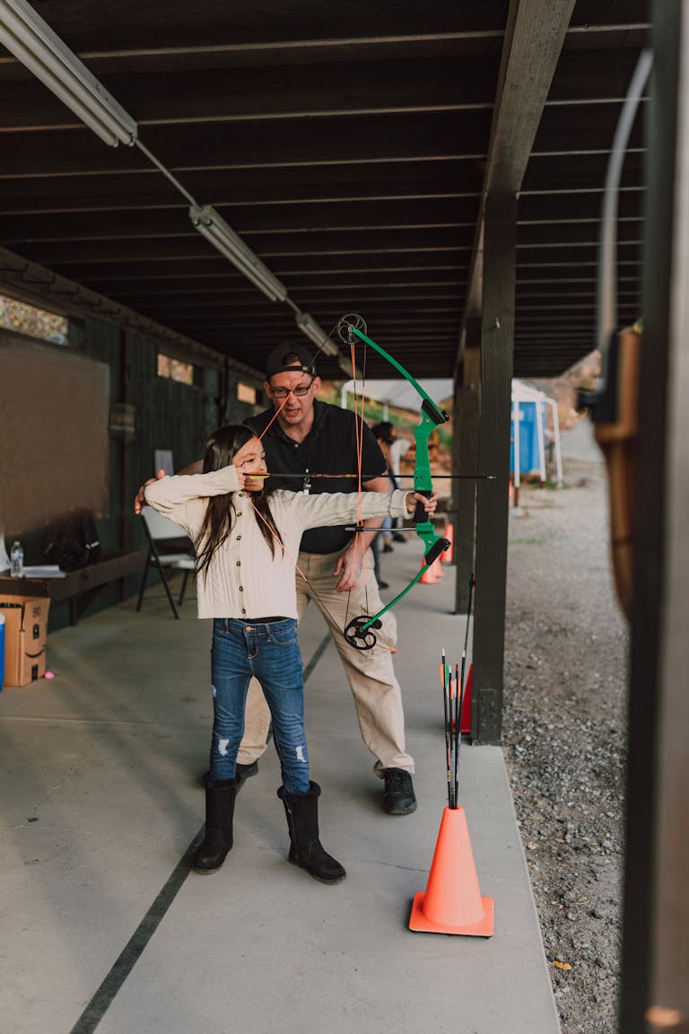 A young girl learns archery under the guidance of an adult at an outdoor range.