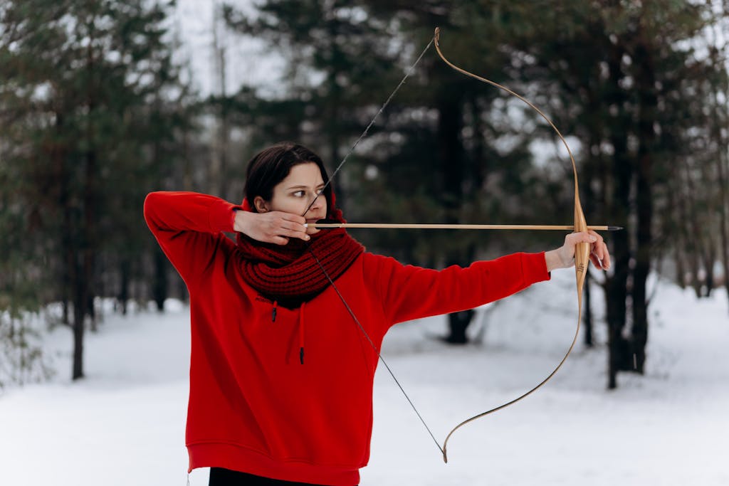 Woman archer in a red hoodie aiming a bow and arrow in a snowy forest during winter.